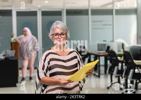 Donna ispanica penosa con capelli grigi in occhiali e abiti casual in piedi con documenti in cartella gialla in ufficio moderno in Costa Rica Foto Stock