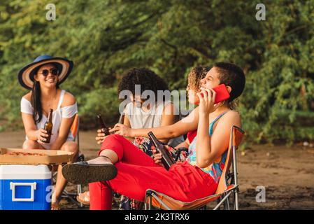 Allegra giovane donna che parla sul telefono cellulare mentre si riunisce con gli amici mangiando pizza e bevendo birra durante il fine settimana estivo sulla spiaggia di Costa Ric Foto Stock