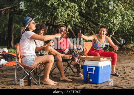 Gruppo di giovani amici allegri seduti sulle sedie e di sedie a sdraio bottiglie di birra mentre si riuniscono sulla spiaggia di sabbia in estate sera in Costa Rica Foto Stock