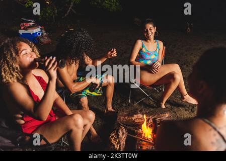 Gruppo di amici etnici allegri con la birra divertirsi mentre si riuniscono intorno a fuoco bruciato durante il luogo di ritrovo estivo in serata sulla spiaggia in Costa R. Foto Stock