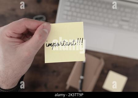 Vista dall'alto verso il basso della mano che tiene il foglietto adesivo giallo con la parola IMPORTANTE sopra la scrivania con il computer portatile e il blocco note Foto Stock