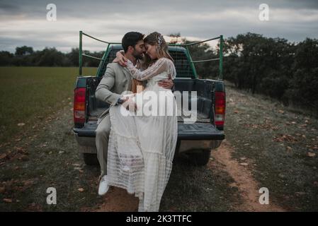 Giovane ragazzo in tuta seduto vicino attraente signora in abito da sposa con gli occhi chiusi che si adagiano sulla spalla maschile e seduto sul pick-up Foto Stock