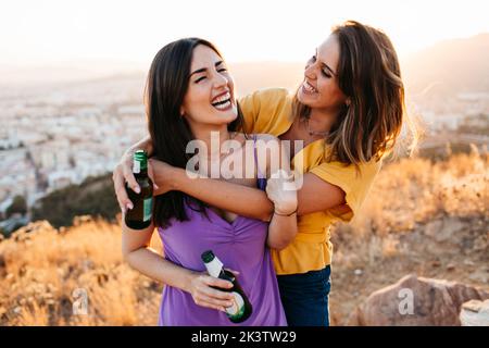 Allegre giovani amiche che si abbracciano in abbigliamento casual tenendo bottiglie di vetro di birra ridere con gli occhi chiusi in piedi sulla cima della montagna Foto Stock