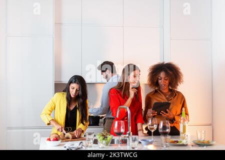 Donna afroamericana che usa il tablet mentre si trova in piedi con gli amici al tavolo con bevande e spuntini e insieme divertirsi e godersi la festa a casa in mod Foto Stock