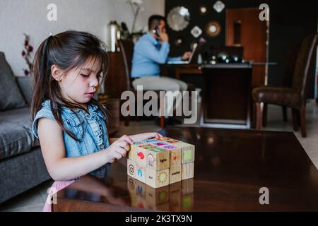 Adorabile bambina etnica giocare con cubi di legno vicino a un padre lavoratore autonomo utilizzando il computer portatile a casa Foto Stock