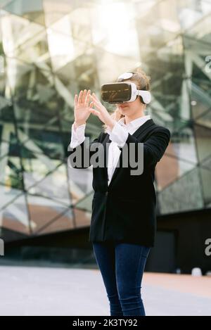 Imprenditrice femminile in stile casual elegante e occhiali VR in piedi nel centro della città e che interagiscono con la realtà virtuale Foto Stock