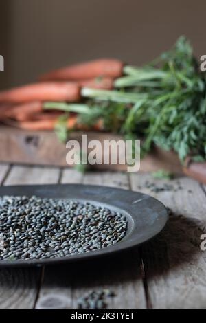 Tavolo in legno con piatto pieno di lenticchie secche su fondo di carote fresche Foto Stock