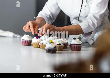 Anonimo Crop femmina panettiere decorazione cupcakes con fiori e frutti di bosco, mentre cucinare sani dessert vegani in panetteria Foto Stock