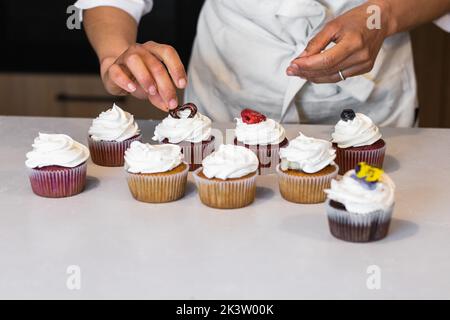 Panettiere femminile corto irriconoscibile in piedi al banco in cucina di panetteria e decorare dolci cupcake vegan con bacche Foto Stock