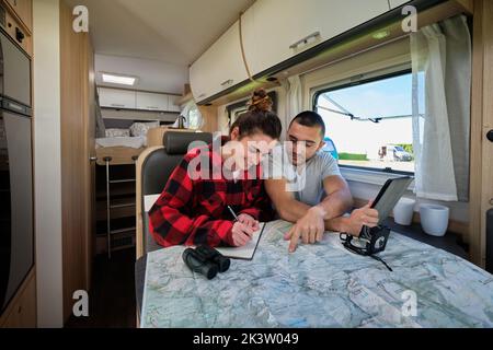 Allegra coppia di viaggiatori che pianificano il percorso di viaggio mentre leggono una mappa cartacea e prendono appunti in un notebook in roulotte Foto Stock