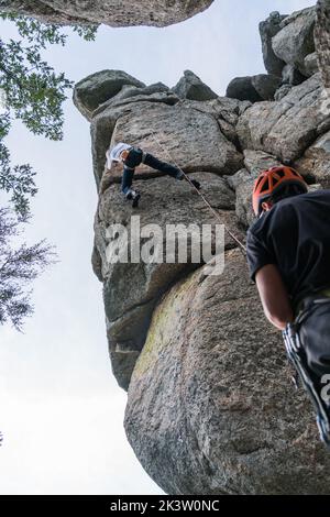 Da sotto arrampicatore in elmetto reggendo la corda e compagno di assicurazione che striscia sulla montagna a strapiombo Foto Stock