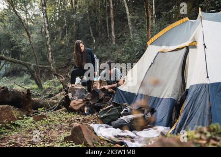 Coppia di escursionisti che fanno falò vicino tenda circondata da alberi che crescono nella foresta mentre esplorano la natura insieme Foto Stock