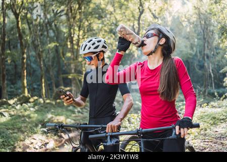 Ciclista femmina acqua potabile contro amico maschio surfing internet sul cellulare durante il viaggio in bicicletta nel bosco Foto Stock