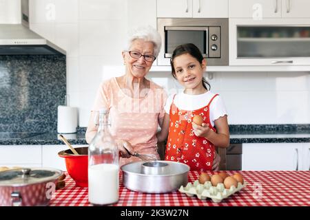 Nonna sorridente in occhiali con frusta abbracciando ragazza affascinante con pin rotolante mentre cucinando e guardando la macchina fotografica in cucina casa Foto Stock