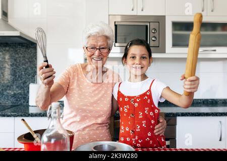 Nonna sorridente in occhiali con frusta abbracciando ragazza affascinante con pin rotolante mentre cucinando e guardando la macchina fotografica in cucina casa Foto Stock