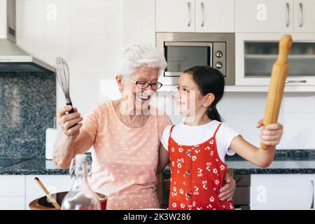 Nonna sorridente in occhiali con frusta abbracciando ragazza affascinante con pin rotolante mentre cucinando e guardando l'un l'altro in cucina casa Foto Stock