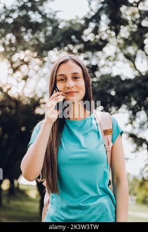 Ragazza studentesca adolescente che indossa una t-shirt acquamarina, facendo una telefonata al suo ragazzo nel parco. Retroilluminazione del sole Foto Stock