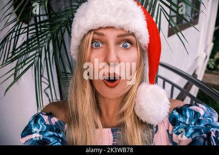 Sorpresa donna eccitata con gli occhi blu che indossano il cappello di santa e l'abito tropicale guarda in macchina fotografica contro lo sfondo di piante verdi. Concetto di Capodanno Foto Stock