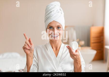 Bella donna di mezza età applicando crema sotto i suoi occhi, facendo il trattamento di bellezza con il prodotto cosmetico naturale Foto Stock