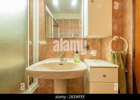 Bagno con specchio senza cornice, lavabo in porcellana bianca con piedistallo coordinato e cabina doccia con accenti bianchi e piastrelle marroni Foto Stock