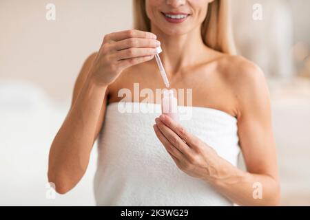 Donna matura che applica siero facciale, facendo la routine di cura della pelle e idratando la pelle, in piedi avvolto in asciugamano Foto Stock