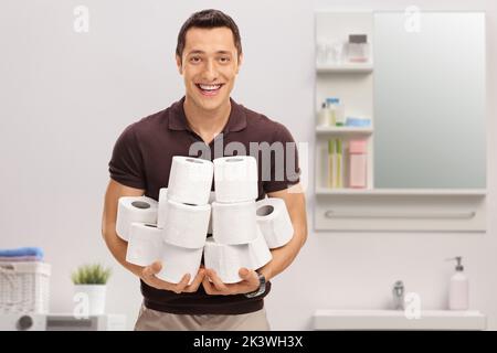 Giovane uomo sorridente che tiene un mucchio di rotoli di carta igienica all'interno del bagno Foto Stock