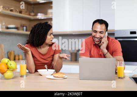 Arrabbiato millenario nero donna trasudare l'uomo, parla al telefono con il computer portatile in cucina interno Foto Stock