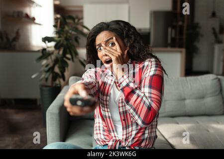 Donna afro-americana impaurita con telecomando siede sul divano e copre il viso con le mani Foto Stock