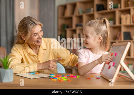 Donna eccitato discorso terapista insegnamento adorabile piccola ragazza alfabeto, facendo parole da lettere di plastica su lavagna bianca Foto Stock