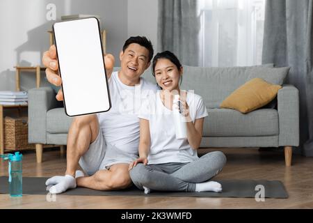 Coppia asiatica sportiva che pratica yoga mattutino a casa, mostrando lo smartphone Foto Stock