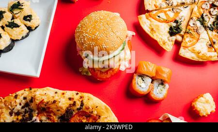 Una vista dall'alto di prelibatezze fast food, tra cui pizza, hamburger e sushi roll su un tavolo rosso Foto Stock