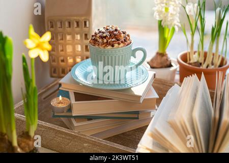 Tazza da caffè con panna montata, fiori primaverili e libro aperto sul davanzale. Concetto caldo e accogliente casa, hygge, lettura libri, riposo e comfort, primavera Foto Stock