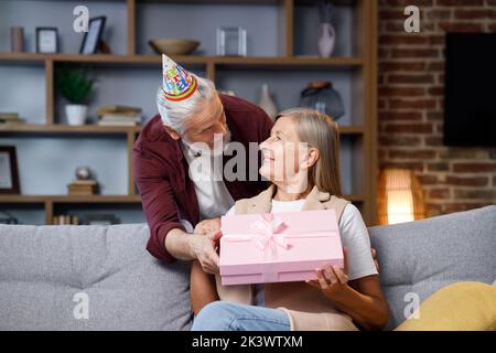 Premuroso vecchio marito dando romantico regalo di compleanno a moglie matura eccitato, amore uomo più anziano sorpresa felice donna di mezza età, scatola rosa aperta, bacio Foto Stock