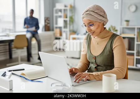 Ritratto minimale di sorridente donna d'affari mediorientale che indossa la fascia mentre scrive al laptop in ufficio, spazio della copia Foto Stock