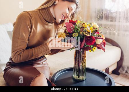 La donna odora il bouquet autunnale fatto di fiori rossi gialli arancioni disposti in vaso a casa. Fiori freschi nei colori autunnali Foto Stock