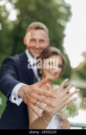 Ritratto di coppia di nozze overjoyed in piedi nel parco in estate, allungando le mani, mostrando anelli d'aggancio dorati. Foto Stock