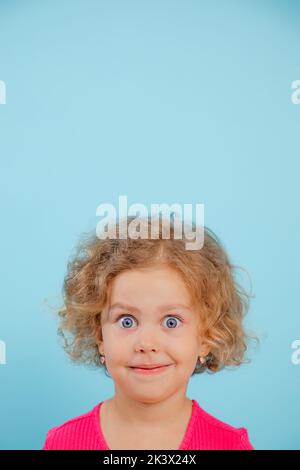 Ritratto di bambina sorridente con capelli ricci, occhi azzurri spalancati che indossano una T-shirt rosa, che guarda su sfondo blu. Foto Stock