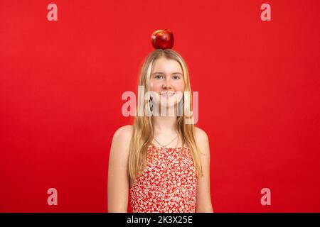 Ritratto di sorridente ragazza adolescente meravigliosa con i capelli giusti che tengono equilibrare la mela rossa grande sulla testa su sfondo rosso. Foto Stock