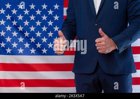 Un uomo d'affari con pollici in segno di vittoria con la bandiera degli Stati Uniti sullo sfondo. Vittoria per gli Stati Uniti. Supporto per la politica statunitense Foto Stock