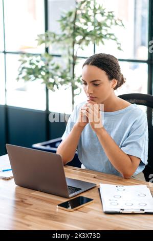 Foto verticale di una donna brasiliana o latina focalizzata di successo, dipendente dell'ufficio, responsabile creativo, siede sul posto di lavoro nell'ufficio moderno, guardando il laptop, pensando a un progetto, progettando Foto Stock