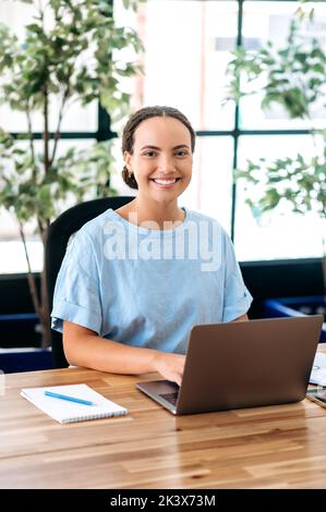 Ritratto verticale di una bella donna d'affari. Sicuro successo latino o giovane donna brasiliana, seo di società, manager creativo, si siede su una scrivania in un ufficio moderno, guardando la fotocamera, sorridendo Foto Stock