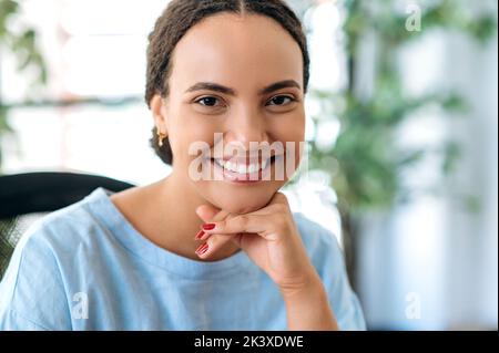 Foto ravvicinata di una bella giovane donna ispanica o brasiliana amichevole, top manager dell'azienda, con una camicia blu, seduta nel suo ufficio, guardando la macchina fotografica, sorridendo felicemente Foto Stock