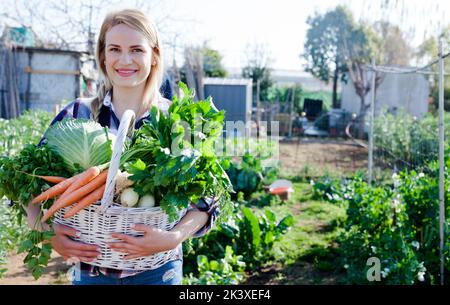 Giovane donna giardiniere che tiene raccolto di verdure e verdi in cestino Foto Stock
