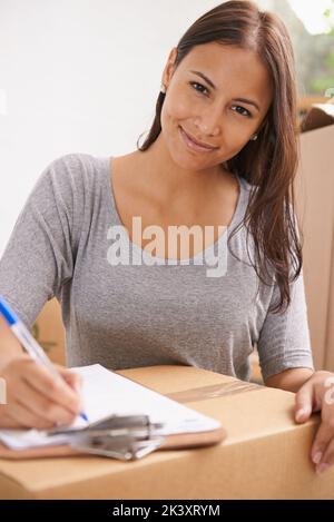 Lasciare iniziare il disimballaggio. Una bella giovane donna sorridente alla macchina fotografica mentre firma un foglio di consegna nella sua nuova casa. Foto Stock