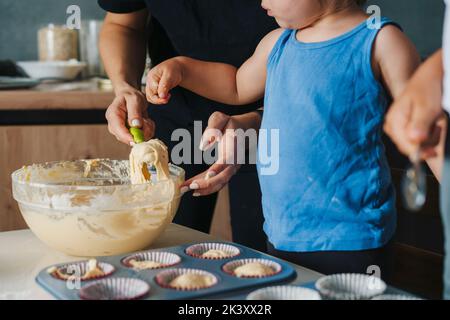 I bambini aiutano la madre a riempire con pasta cruda gli stampi in silicone per cupcake. Cibo dolce. Processo di cottura. Versare l'impasto per muffin negli stampi Foto Stock