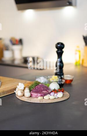 Bistecca di manzo crudo wrap e ingredienti di varietà su piatto di vimini circolare con salsa, sale e burro in mini recipiente di vetro sul banco da cucina in granito nero surf Foto Stock