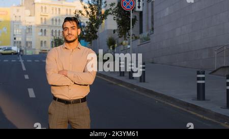 Serio sicuro riuscito ragazzo elegante ispanico arabo indiano business uomo si trova sulla strada in città attraverso le armi meditando piano strategia di pensiero Foto Stock