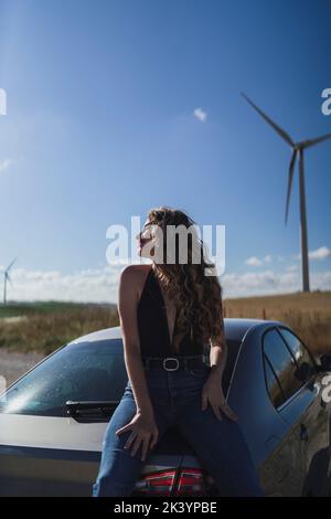 Una foto verticale di una bella donna caucasica che si posa vicino ad un'auto in un campo con una turbina eolica Foto Stock