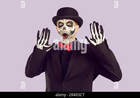 Giovane uomo con trucco scheletro e in elegante vestito Halloween solleva le mani con paura. Foto Stock
