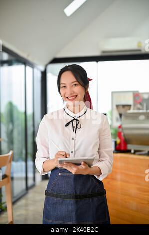 Ritratto, attraente e affascinante giovane asiatica ragazza coffee shop proprietario o cameriera si trova di fronte al banco cassa con il suo tablet. Piccolo busin Foto Stock